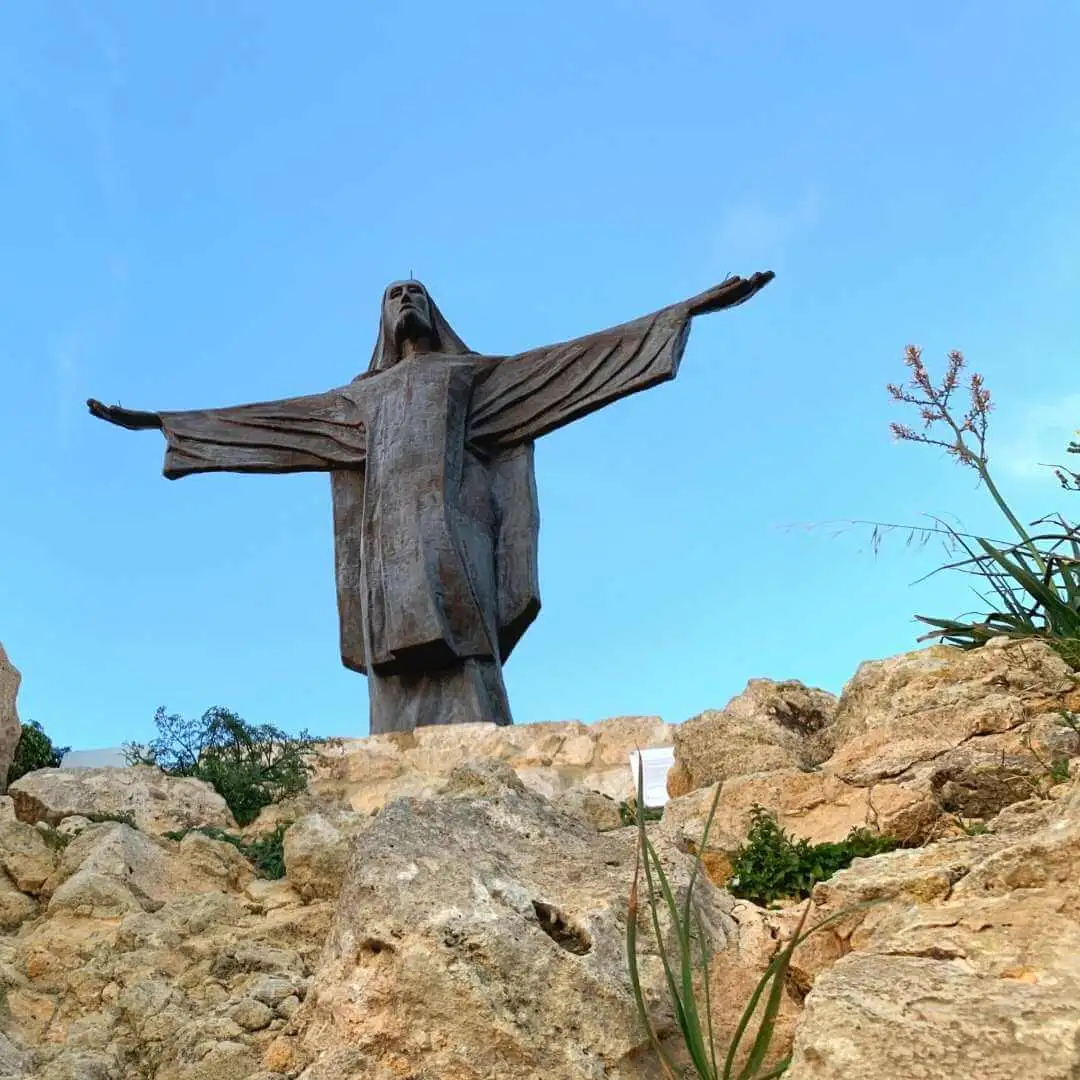 A close-up, low-angle view of the Tas-Salvatur (Risen Christ) statue standing on a rocky hilltop against a blue sky.