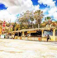 The colorful yellow beach kiosk with a thatched roof at Hondoq Bay in Qala, serving food and drinks under palm trees.