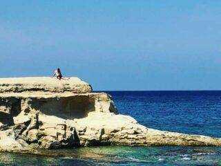 A lone sunbather relaxing on the flat limestone rock formations along the coast of Marsalforn, overlooking the deep blue sea.