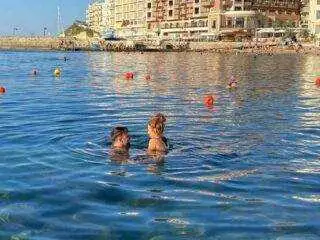 A couple swimming in the calm waters of Marsalforn Bay during golden hour with seaside hotels and buildings in the distance.