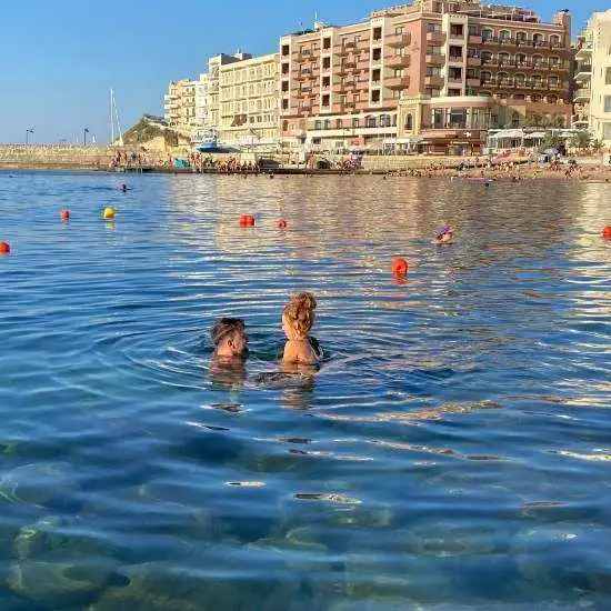 A couple swimming in the calm waters of Marsalforn Bay during golden hour with seaside hotels and buildings in the distance.