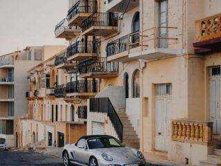 A silver Porsche convertible parked on a sloped street in Marsalforn lined with traditional limestone Maltese balconies.