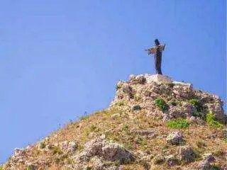 The iconic Tas-Salvatur statue (Risen Christ) standing atop the hill overlooking the Marsalforn valley against a clear blue sky.
