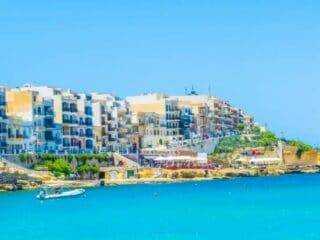 A bright, sunny view of the Marsalforn promenade featuring colorful apartment buildings and the clear blue Mediterranean sea.