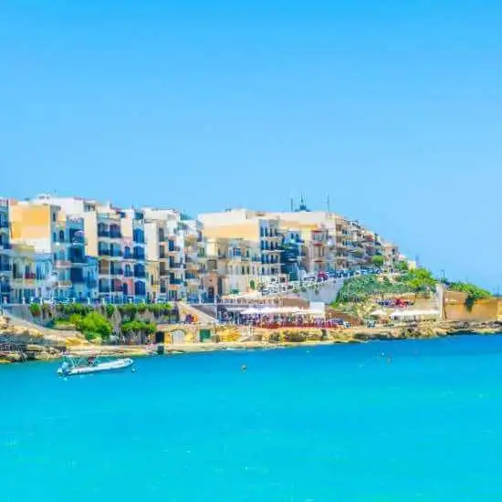A bright, sunny view of the Marsalforn promenade featuring colorful apartment buildings and the clear blue Mediterranean sea.