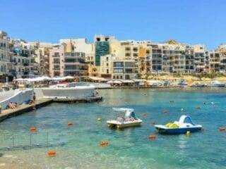 The main beach area in Marsalforn with rental paddle boats floating in the turquoise water and the town center in the background.