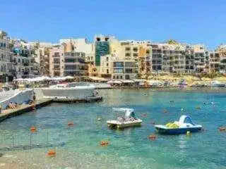 The main beach area in Marsalforn with rental paddle boats floating in the turquoise water and the town center in the background.