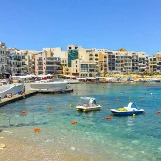 The main beach area in Marsalforn with rental paddle boats floating in the turquoise water and the town center in the background.