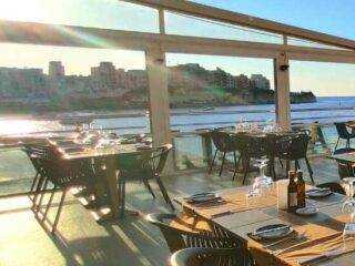 Interior of a waterfront restaurant in Marsalforn, Gozo, with tables set for dinner and a sunset view over the bay.
