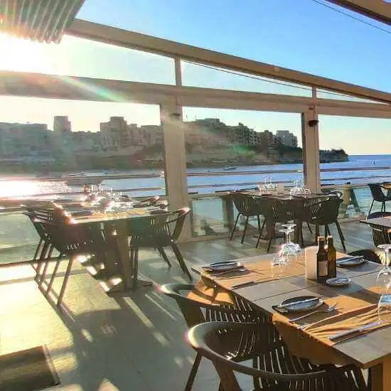 Interior of a waterfront restaurant in Marsalforn, Gozo, with tables set for dinner and a sunset view over the bay.