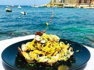 A plate of fresh spaghetti alle vongole (clam pasta) served at a seaside restaurant with the Marsalforn bay visible in the background.