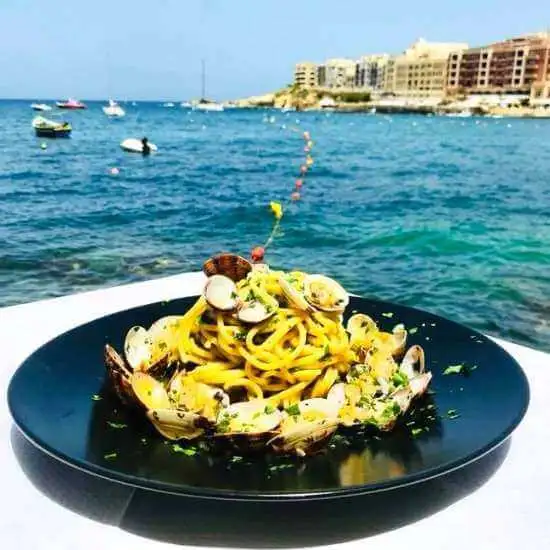 A plate of fresh spaghetti alle vongole (clam pasta) served at a seaside restaurant with the Marsalforn bay visible in the background.