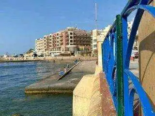 A view along the Marsalforn breakwater featuring a blue metal railing in the foreground and seafront hotels in the background.