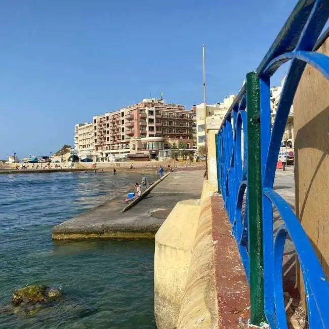 A view along the Marsalforn breakwater featuring a blue metal railing in the foreground and seafront hotels in the background.