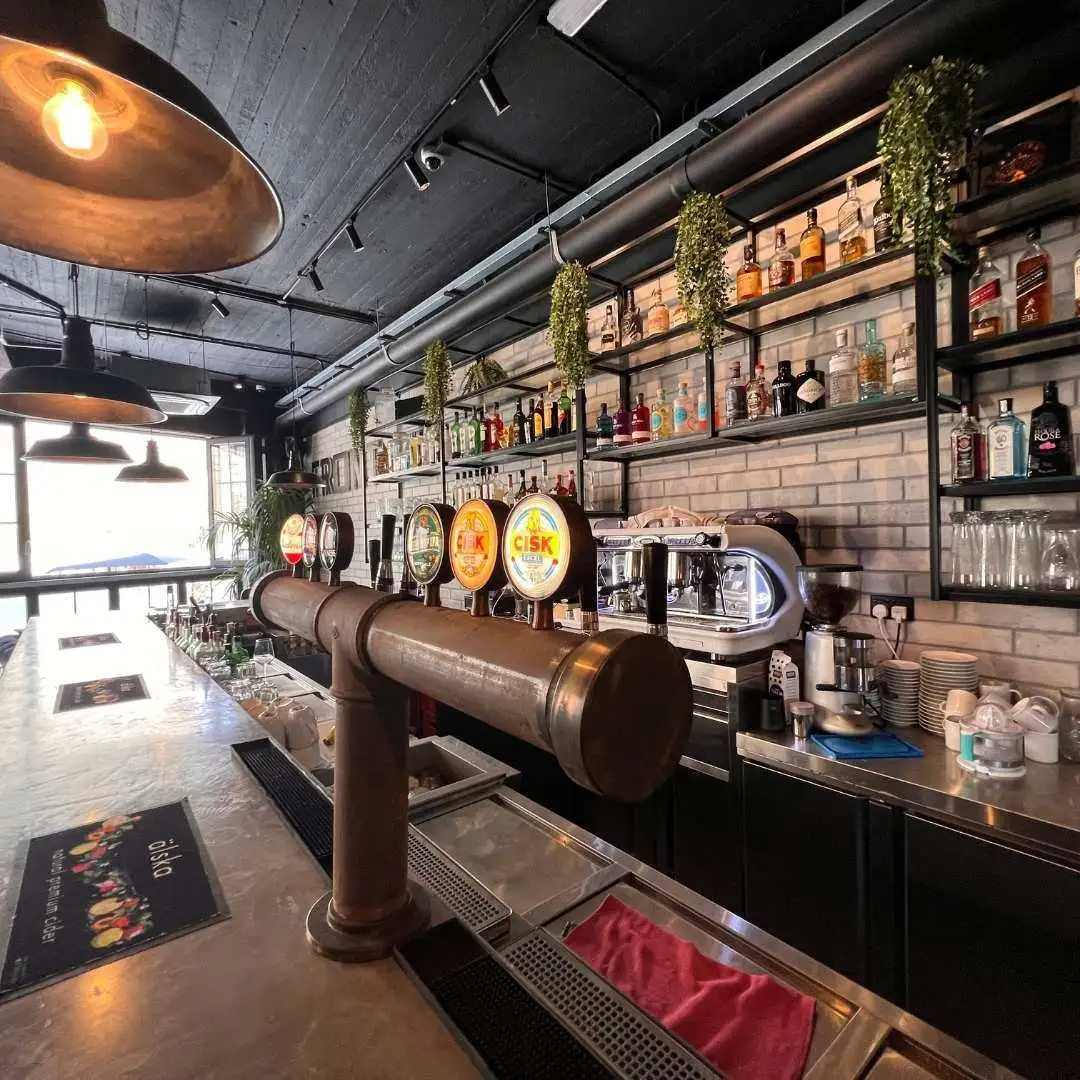 The interior of a modern bar in Qala featuring a long counter with Cisk beer taps, industrial lighting, and shelves stocked with spirits.