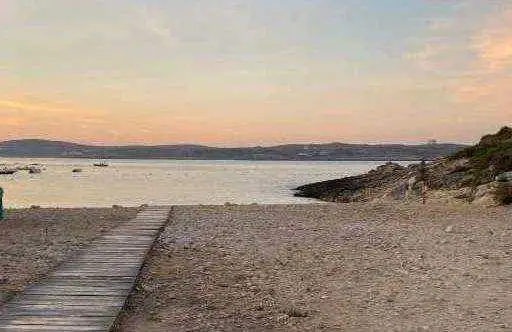 A wooden boardwalk leading down to the pebbly beach in Marsalforn Bay during a peaceful sunset, with calm waters in the background.