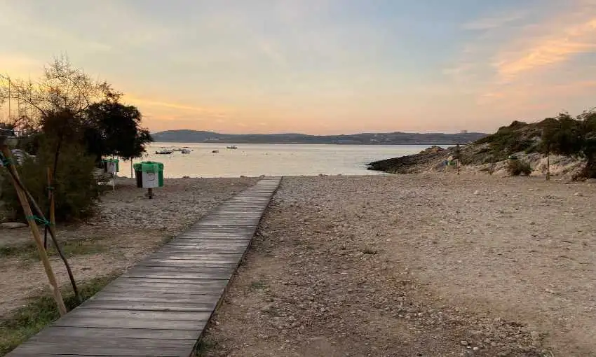 A wooden boardwalk leading down to a pebbly beach during a peaceful golden hour, perfect for a sunrise walk in Qala.