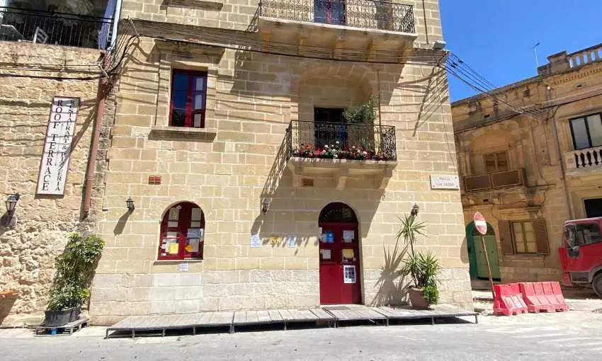 The traditional limestone facade of a restaurant and pizzeria in Marsalforn, featuring a red door and a sign for a roof terrace.