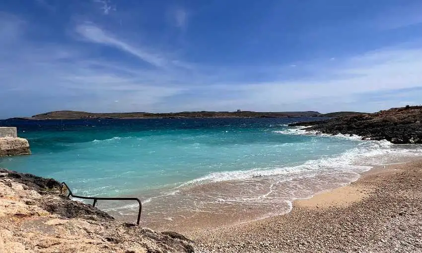 The crystal clear turquoise waters and pebbly shore of Hondoq ir-Rummien beach in Qala, with a view of Comino island in the background.
