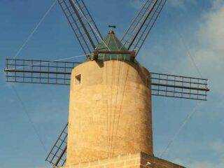 A low-angle view of the historic windmill in Qala, showcasing its limestone structure and wooden vanes against a blue sky.