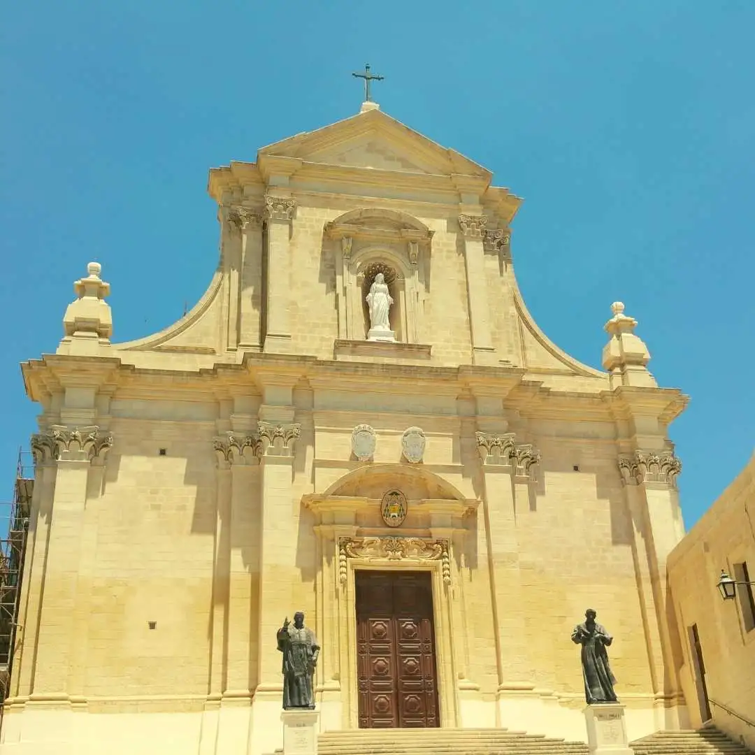 The sunlit Baroque facade of the Parish Church of St. Joseph in Qala, Gozo, featuring two statues at the entrance and a clear blue sky.