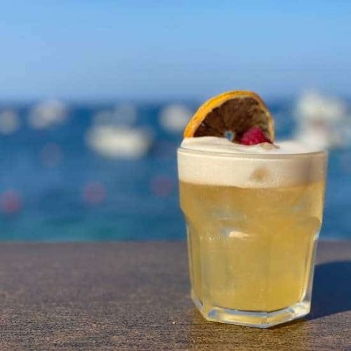 A refreshing cocktail garnished with dried citrus and raspberry, set against a blurred background of the blue Mediterranean sea in Marsalforn.