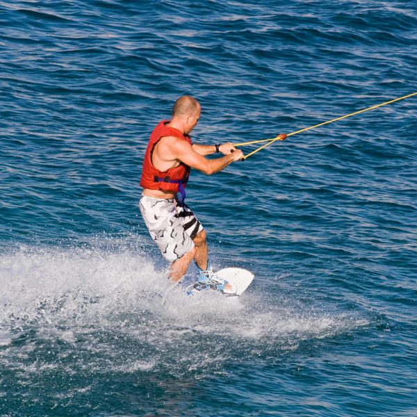 A man wakeboarding across the deep blue sea in Marsalforn, wearing a red life jacket and creating a splash.